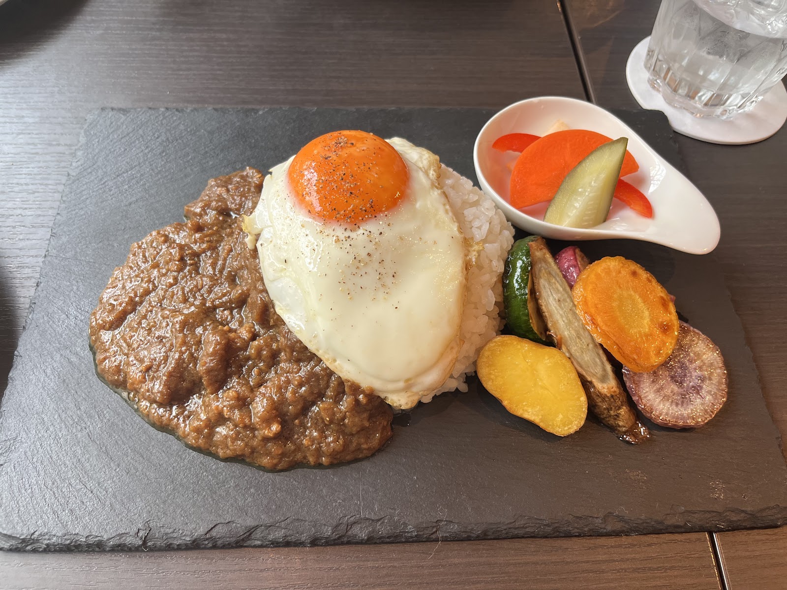 スレートの皿に、ひき肉のカレーとご飯、その上に目玉焼き。焼いた根菜を添え、白い小皿にピクルス。昼の光。