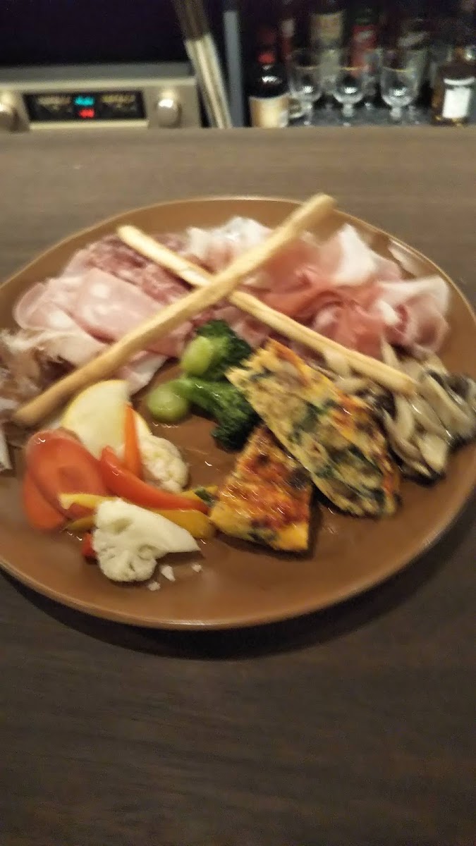 茶色い丸皿に、薄切りの肉、ゆでた緑の野菜、赤と黄のパプリカ、卵を使った四角い料理、細長いパンが二本。奥はバーの棚。暗く、ピントのやわらかい一枚。
